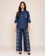 A woman modeling a matching navy blue two-piece set consisting of a short-sleeve button-up shirt and wide-leg trousers, both featuring white floral embroidery.