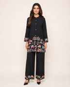 A woman modeling a black two-piece kurta and trouser set with colorful embroidered animal motifs around the hem and cuffs, photographed against a plain light beige studio background.