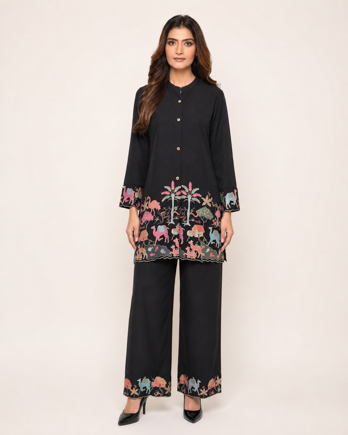 A woman modeling a black two-piece kurta and trouser set with colorful embroidered animal motifs around the hem and cuffs, photographed against a plain light beige studio background.