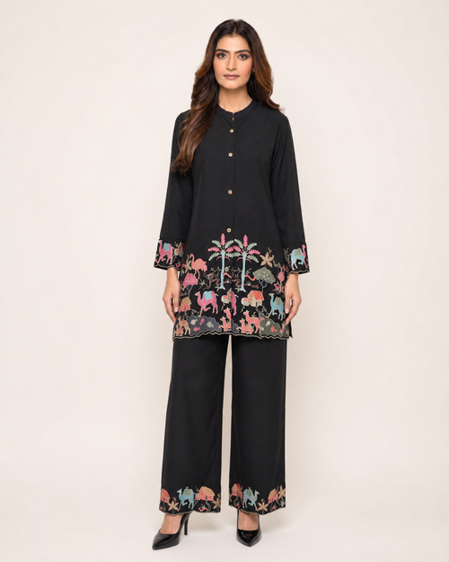A woman modeling a black two-piece kurta and trouser set with colorful embroidered animal motifs around the hem and cuffs, photographed against a plain light beige studio background.