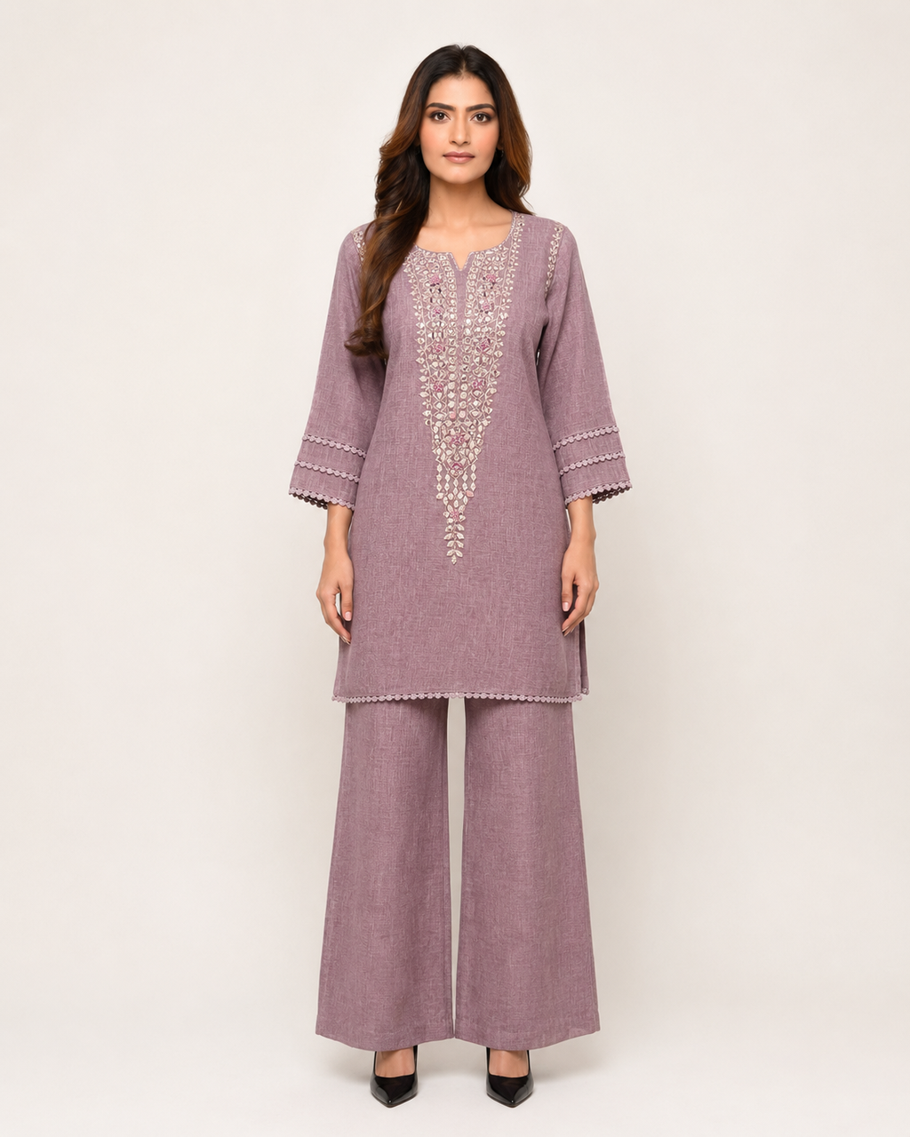 A woman modeling a dusty purple two-piece kurta and trouser set with white embroidered details on the chest and sleeve cuffs, set against a plain light beige background.