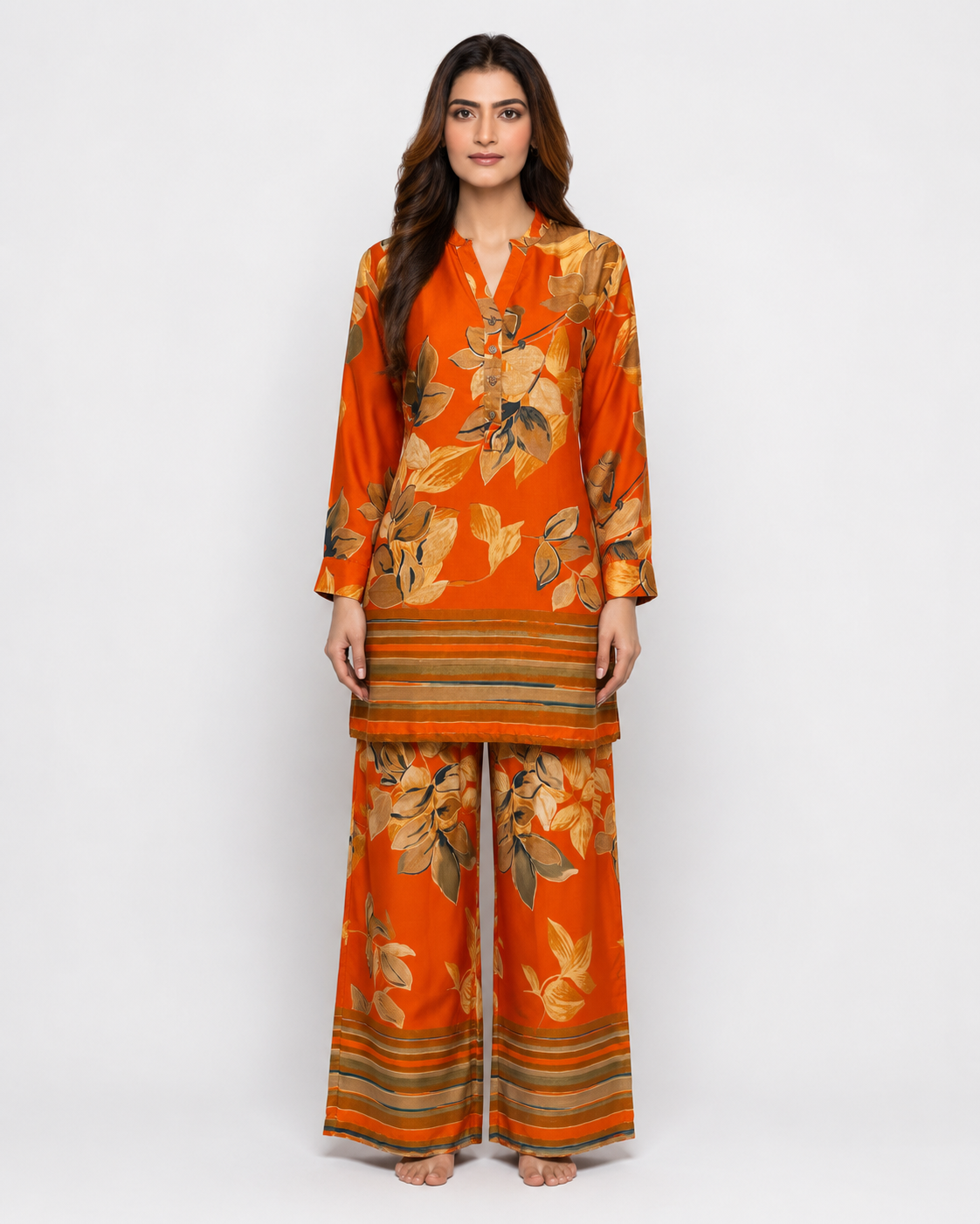 A woman modeling a two-piece orange kurta and trouser set with a floral print and striped borders, photographed against a plain white studio background.