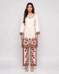 A woman modeling a traditional three-piece salwar kameez suit in cream with vibrant red, pink, and green floral embroidery. The set includes a long-sleeve tunic, matching trousers, and a coordinating belt, shown against a plain light gray studio background.