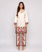 A woman modeling a traditional three-piece salwar kameez suit in cream with vibrant red, pink, and green floral embroidery. The set includes a long-sleeve tunic, matching trousers, and a coordinating belt, shown against a plain light gray studio background.