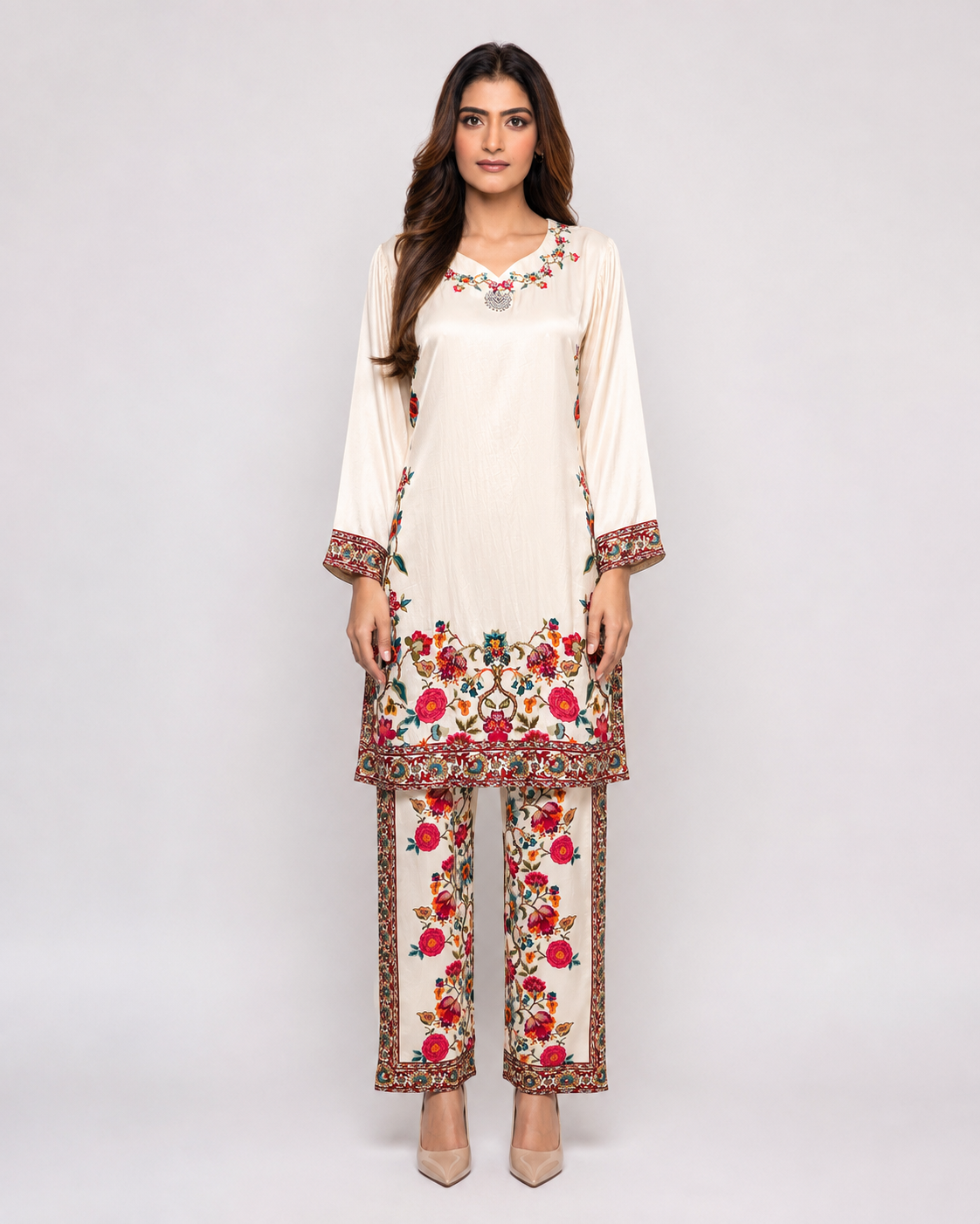 A woman modeling a traditional three-piece salwar kameez suit in cream with vibrant red, pink, and green floral embroidery. The set includes a long-sleeve tunic, matching trousers, and a coordinating belt, shown against a plain light gray studio background.