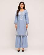 A woman modeling a light blue three-piece traditional outfit consisting of a long-sleeve embroidered kurta, matching wide-leg trousers, and black pointed-toe heels against a plain light beige studio background.