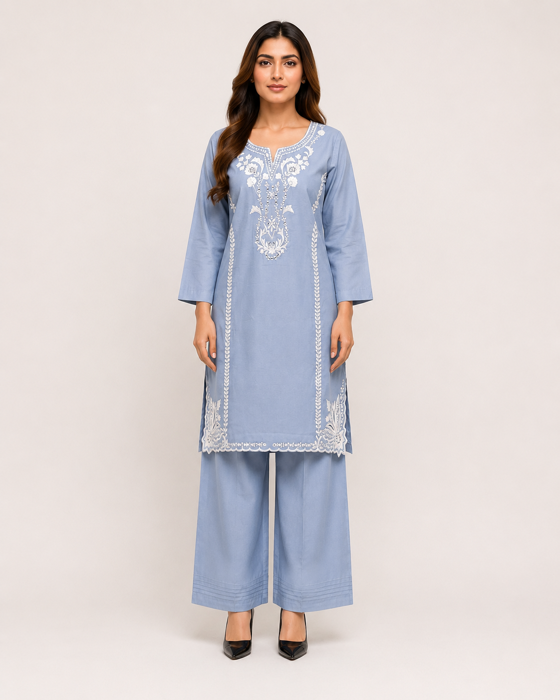 A woman modeling a light blue three-piece traditional outfit consisting of a long-sleeve embroidered kurta, matching wide-leg trousers, and black pointed-toe heels against a plain light beige studio background.