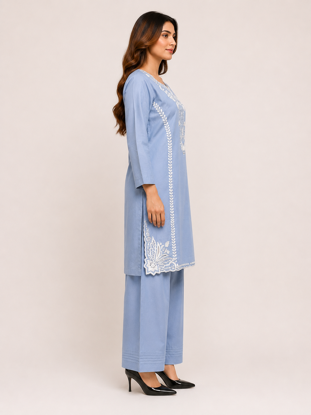 A woman modeling a light blue three-piece kurta set with white embroidery, consisting of a long tunic, matching pants, and black high heels, set against a plain beige studio background.