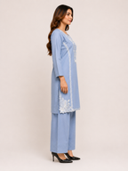 A woman modeling a light blue three-piece kurta set with white embroidery, consisting of a long tunic, matching pants, and black high heels, set against a plain beige studio background.