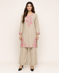 A woman modeling a beige three-piece kurta set with pink floral embroidery, consisting of a long-sleeve tunic, matching wide-leg pants, and black pointed-toe heels, against a plain light beige studio background.