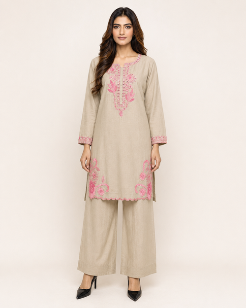 A woman modeling a beige three-piece kurta set with pink floral embroidery, consisting of a long-sleeve tunic, matching wide-leg pants, and black pointed-toe heels, against a plain light beige studio background.