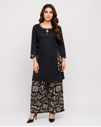 A woman modeling a black kurta with silver embellishments and wide-leg black pants featuring a beige and gold block-print pattern, set against a plain light gray background.
